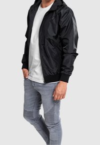 Svart vindjacka med huva, dragkedjestängning och sidfickor; matchad med ljusgrå skinny jeans med texturerade paneldetaljer.