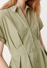 Olivově zelené šaty košilového střihu s krátkými rukávy, knoflíkovým zapínáním vpředu a nařaseným pasem. Hladká tkanina s volným střihem a jemnou texturou.