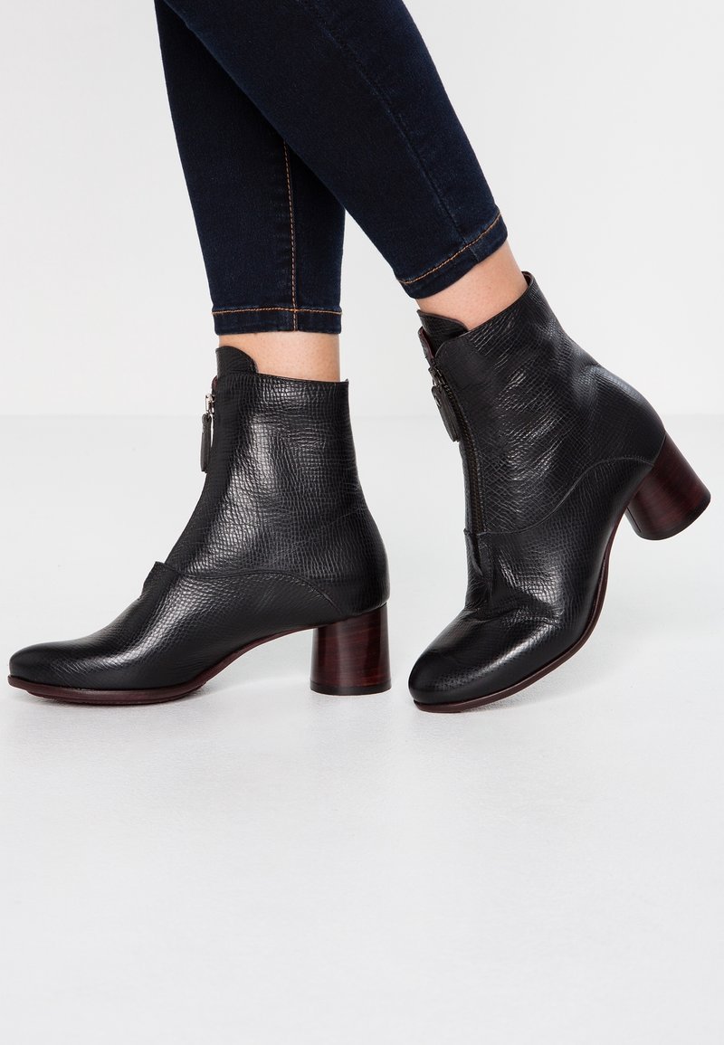 Černé texturované kožené kotníkové boty se zipy na boku a blokovými podpatky, nošené s tmavými úzkými jeansy na světlé podlaze.