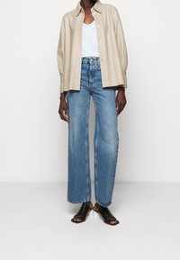 Béžová kožená košile-jacket přes bílý top na ramínkách, doplněná modrými kalhotami s širokými nohavicemi a černými sandály na podpatku. Hladké textury a uvolněný střih.