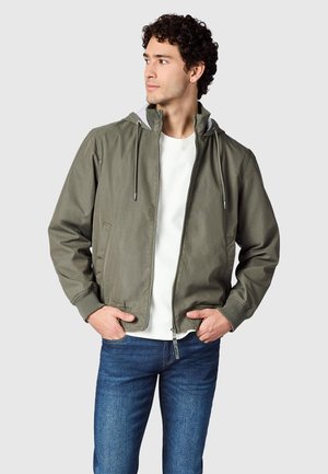 Nuori mies, jolla oliivi vihreä hupullinen takki, valkoinen paita ja siniset farkut, katsoo vasemmalle kädet takin taskuissa.