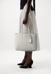 Borsa tote in pelle bianca con manici lunghi, zip superiore e una piccola etichetta. Texture liscia e design minimalista su uno sfondo neutro.