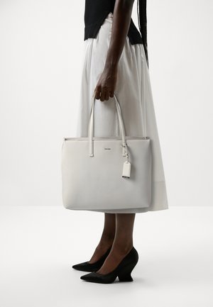 Borsa tote in pelle bianca con manici lunghi, zip superiore e una piccola etichetta. Texture liscia e design minimalista su uno sfondo neutro.
