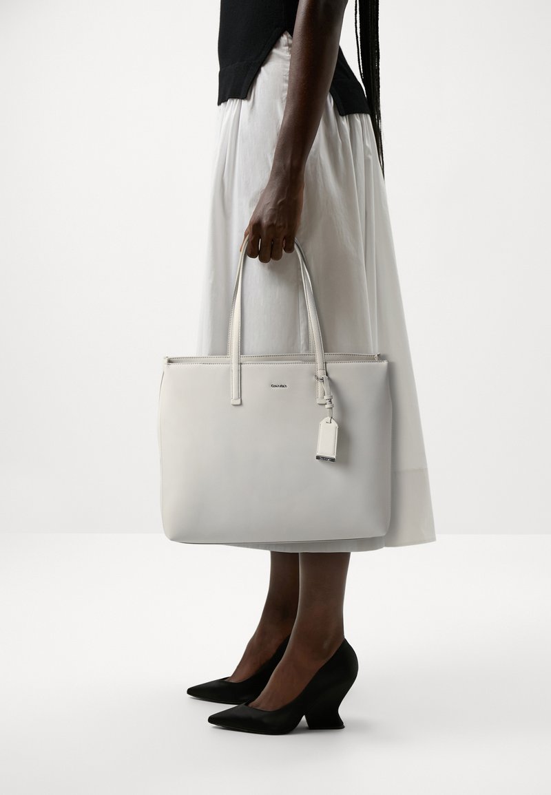 Borsa tote in pelle bianca con manici lunghi, zip superiore e una piccola etichetta. Texture liscia e design minimalista su uno sfondo neutro.