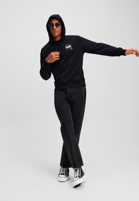 Hanorac negru cu buzunar frontal și un logo pe piept, asortat cu blugi închisi și adidași înalți, creând un look casual.