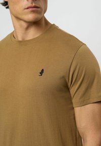 Hnědá bavlněná tričko s kulatým výstřihem, krátkými rukávy a malým černým vyšitým logem na levé hrudi. Hladká textura, volný střih.