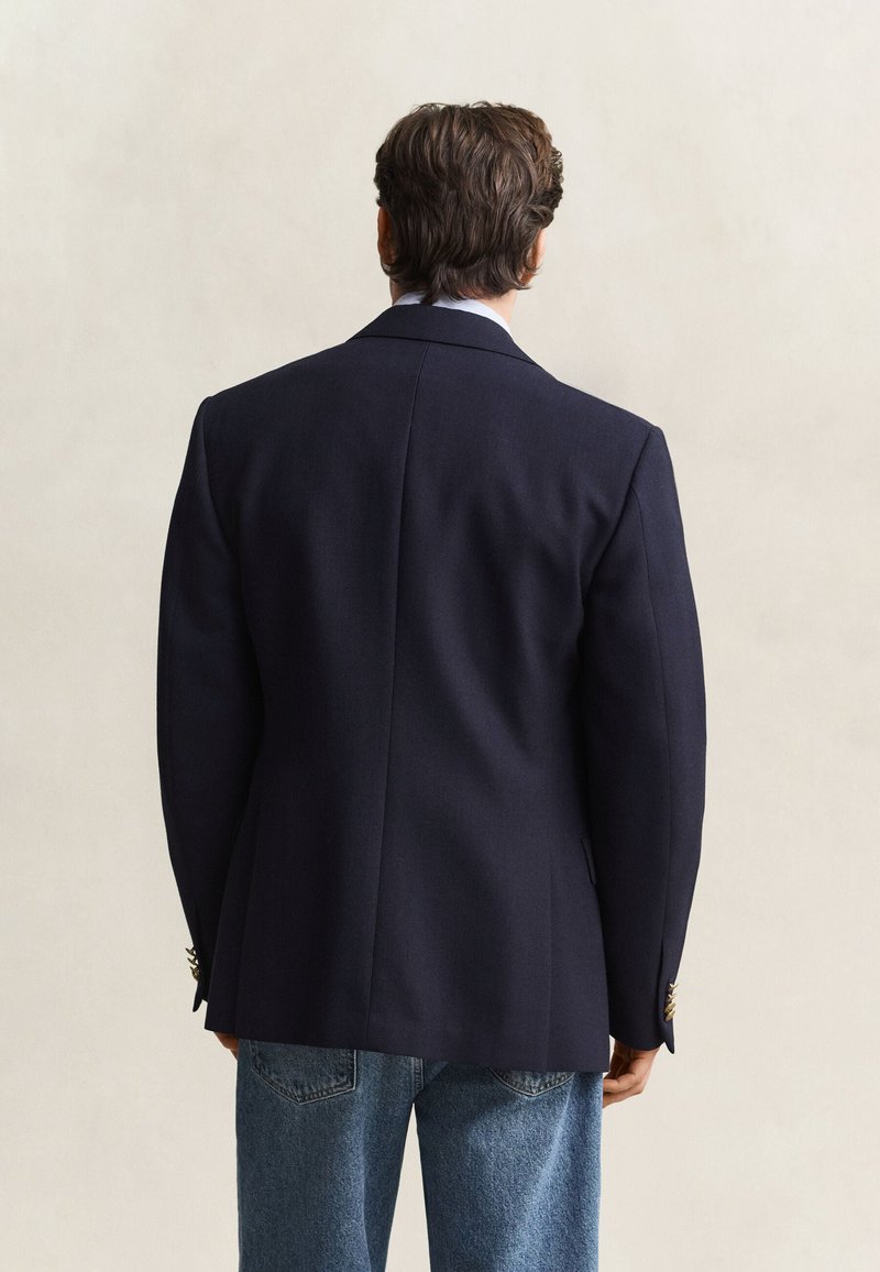 Mand iført en mørk navy blazer og blå jeans, der står med ryggen mod kameraet mod en ensfarvet lys baggrund.