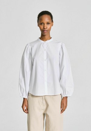 Kvinde iført en hvid, knaplukket bluse med puffede lange ærmer og beige bukser, stående mod en ensfarvet lys baggrund.