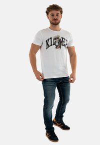 T-shirt blanche à manches courtes présentant un graphique d'ours avec une batte, associée à un jean bleu et des baskets noires avec des accents orange.