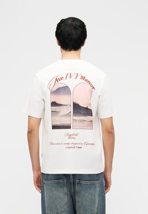 Homme portant un t-shirt blanc avec une impression pittoresque rose et violette représentant des vagues et des collines, avec le texte « The IVY Manor » et « Bayfield Harbor » au dos.