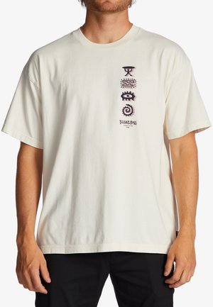 Mies, joka pukeutunut valkoiseen t-paitaan, jossa on neljä abstraktia mustaa ja vaaleanpunaista symbolia pinottuna pystysuunnassa vasemman yläreunan rinnan kohdalla pienen tekstin yläpuolella.