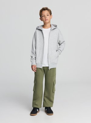 Szara bluza z kapturem na zamek, pod spodem biała koszulka, oliwkowe spodnie cargo z bocznymi kieszeniami oraz czarne trampki z gumową podeszwą.