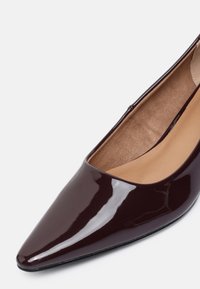 Chaussure plate en cuir verni marron foncé à bout pointu avec semelle intérieure beige rembourrée sur fond blanc.