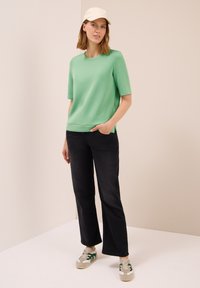Femme portant un haut à manches courtes vert clair, un pantalon noir straight-leg, des baskets beiges avec des rayures vertes, et une casquette blanche, se tenant dans une pièce neutre.