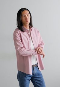 Lys rosa langermet skjorte med knapper og krage, parret med en hvit T-skjorte og blå mønstrede jeans. Glatt stofftekstur.
