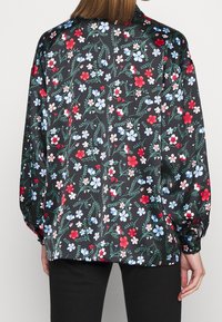 Blusa floreale con sfondo nero, caratterizzata da fiori colorati in rosso, blu e verde. Maniche lunghe con polsini arricciati. Tessuto di texture liscia.