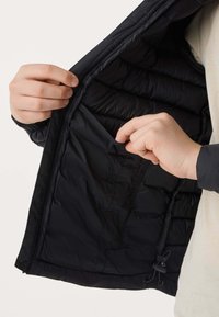 Des mains ouvrant la veste noire matelassée pour révéler et accéder à une poche intérieure à la poitrine avec un petit cordon de serrage à l'ourlet.