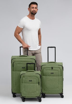 Drie groene polyester koffers in verschillende maten met gladde texturen, zwarte ritsen en draaiwielen, vastgehouden door een persoon.
