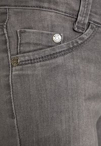 Tejido de mezclilla gris con un bolsillo frontal con herrajes metálicos marcados como "BLUE" y "EFFECT". Apariencia sutilmente texturizada.