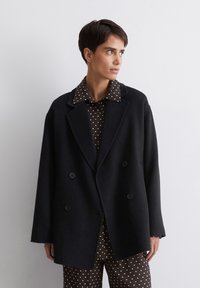 Zwarte dubbelgeknoopte jas van zwaar materiaal, met brede revers en twee voorknopen, gedragen over een bruine blouse met stippen.