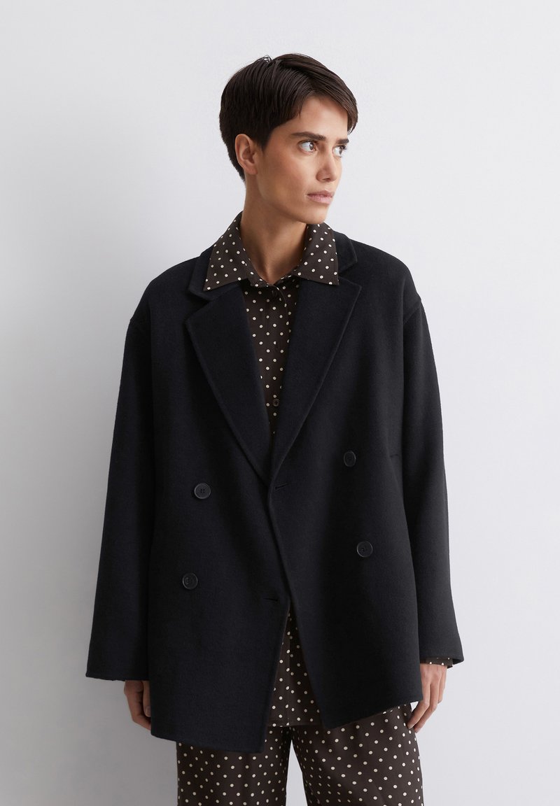 Zwarte dubbelgeknoopte jas van zwaar materiaal, met brede revers en twee voorknopen, gedragen over een bruine blouse met stippen.