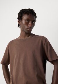 Young Black man with short twists wearing a loose brown t-shirt, standing against a plain white background with hands on hips.