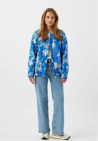 Blå blomsterjakke med hvide mønstre, let stof, afslappet pasform, lynlås lukning, parret med lyseblå jeans og sneakers.
