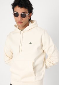 Hoodie couleur crème avec une poche kangourou et des poignets côtelés. Présente un petit logo vert Lacoste sur la poitrine gauche. Texture de tissu lisse.