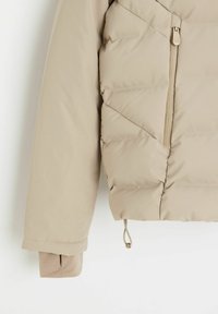 Beżowa kurtka puchowa z teksturowanym materiałem, ukośnym pikowaniem, kieszenią na zamek oraz regulowanym sznurkiem na dole i mankietach.