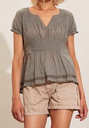 Kvinde iført en kortærmet grå bluse med smock-detaljer og beige cargoshorts med store forlommer.