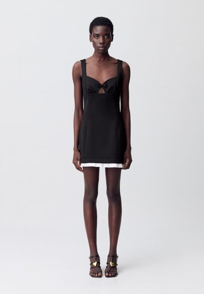 Vestido negro con corsé ajustado, acento de lazo y detalle de corte. El dobladillo presenta un ribete plisado blanco. La modelo lleva tacones negros con tiras.