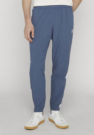 Uomo che indossa pantaloni da jogging con vita elastica blu e sneakers bianche in piedi davanti a uno sfondo semplice.