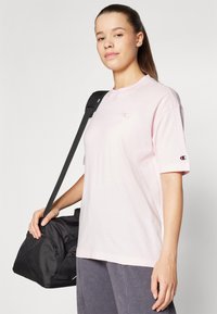 Lyserød bomulds t-shirt med korte ærmer, der har et lille broderet logo. Modellen holder en sort duffeltaske med skulderrem.