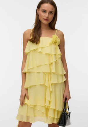 Mujer con vestido amarillo sin mangas de varias capas con detalle floral en el hombro, sosteniendo un pequeño bolso negro sobre un fondo liso.