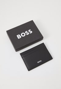 Musta nahkainen korttikotelo, jossa on useita lokeroita ja valkoinen "BOSS" -logo. Mukana musta laatikko valkoisella painatuksella. Silkinpehmeä pinta.