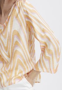 Donna che indossa una blusa a maniche lunghe con strisce ondulate arancioni, rosa e bianche, con la mano poggiata nella tasca di pantaloni bianchi.