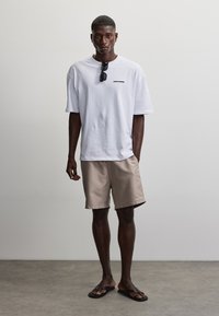 Jeune homme debout portant un T-shirt blanc oversize, un short beige, des tongs noires et des lunettes de soleil accrochées au col du T-shirt sur un fond uni.