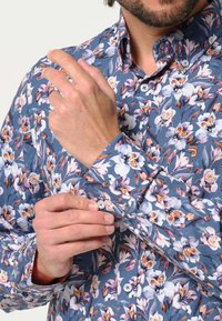 Blauwe bloemenhemel met witte en oranje bloempatronen, met knoopsluiting aan de voorkant en mouwdetails. De stof lijkt glad met een medium fit.