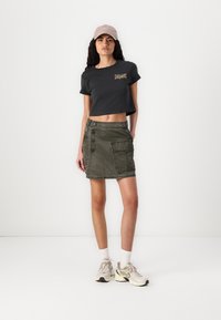 T-shirt grigio corto con dettagli ricamati colorati, abbinato a una gonna in denim khaki con bottoni e tasca laterale. Sneakers neutre completano l'outfit.