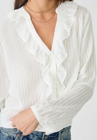 Vit button-up blus med vertikala ränder och volangdetaljer längs halsringningen. Tillverkad av ett lätt, texturerat tyg.