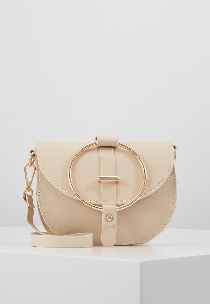 Beige läder crossbody-väska med en rund klaff, gyllene cirkulär detalj, justerbar rem och mjuk textur. Stängs med en matchande rem.