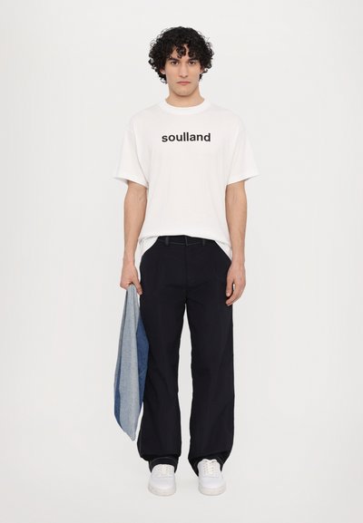 White cotton t-shirt with "soulland" printed in black, paired with black pants and white sneakers. A blue denim jacket is held in the left hand.
