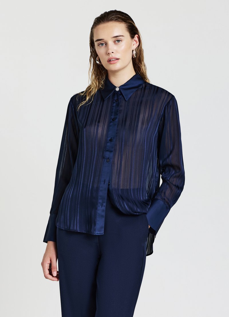 Marinebluse mit halbdurchsichtigen Streifen, klassischem Kragen und einem dekorativen Knopf am Ausschnitt. Lange Ärmel.