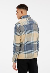 Chemise à carreaux avec un mélange de couleurs beige, bleu et gris. Texture douce, col boutonné et coupe décontractée. Vue de dos montrée.