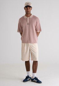 Red and white patterned polo shirt, beige shorts, navy sneakers with white stripes, and a cream cap with a logo, standing on a neutral background.