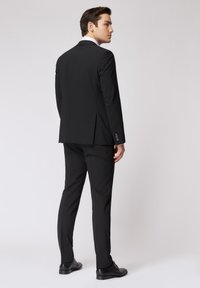 Completo nero con giacca su misura e pantaloni abbinati. Tessuto liscio, rever affilati e dettagli dei bottoni visibili sulle maniche.