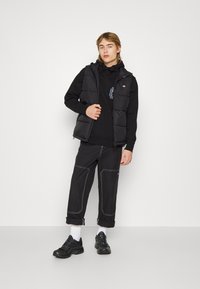 Gilet imbottito nero sopra una felpa nera con cappuccio, abbinato a pantaloni larghi neri con cuciture bianche e sneakers nere con calzini bianchi.