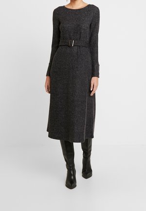 Kobieta ubrana w szarą, długorękawową sukienkę midi z paskiem oraz czarne kozaki do kolan na jednolitym jasnym tle.