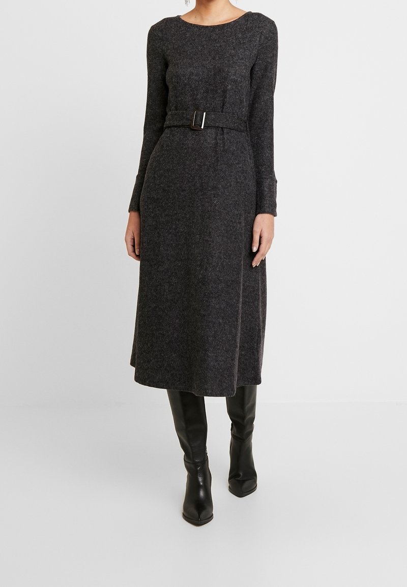 Femme portant une robe midi ceinturée à manches longues de couleur gris foncé avec des bottes noires montantes jusqu'aux genoux, sur un fond clair uni.