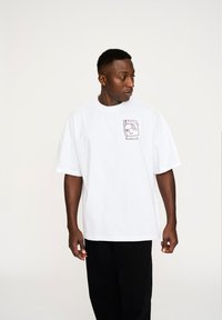 Hvid bomulds t-shirt med en afslappet pasform, prydet med et lilla grafisk design på venstre brystområde. Bæres med sorte bukser.